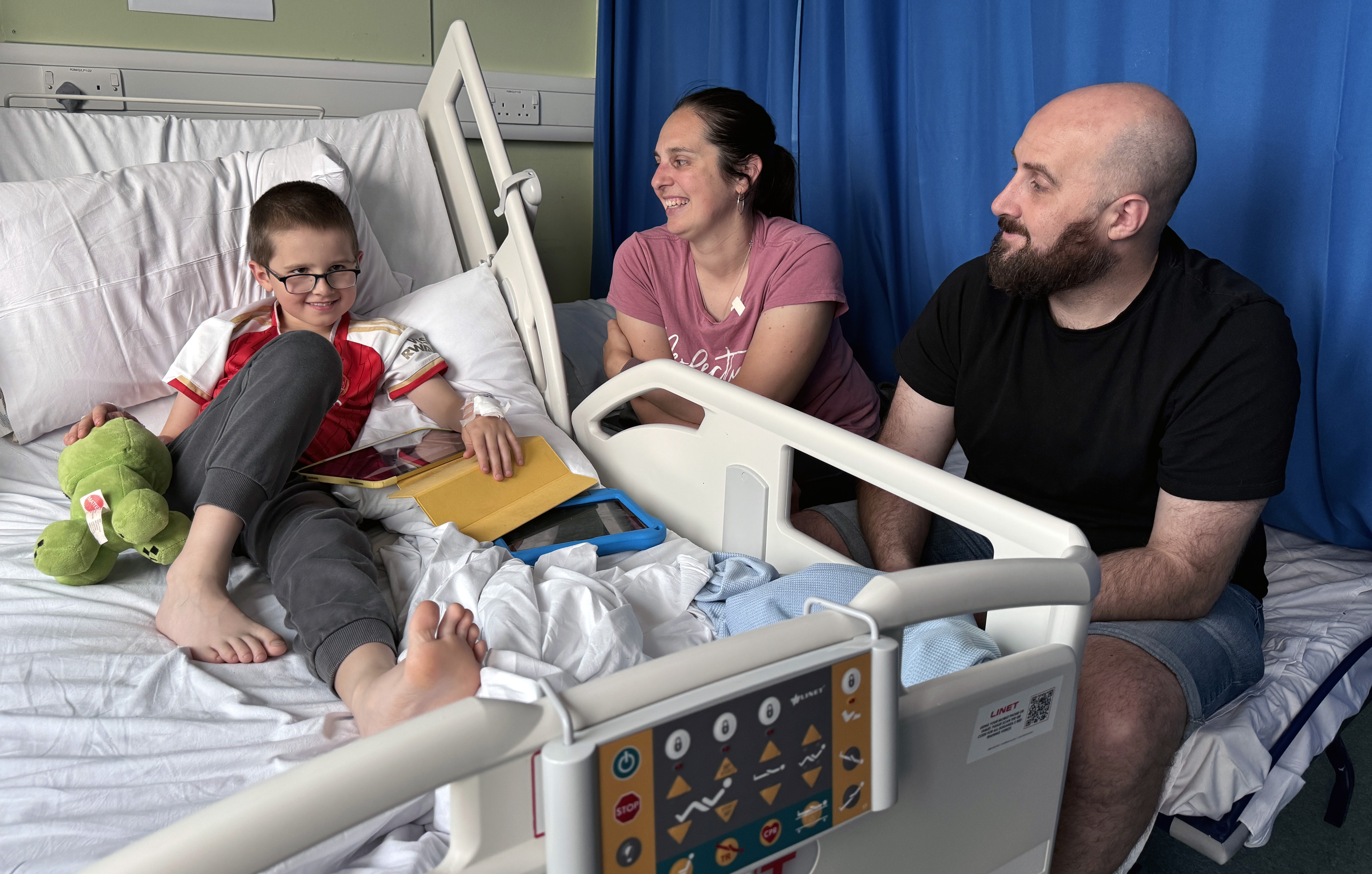 Reece Wilton smiling sat on a hospital bed, with his mum and dad next to him also smiling.