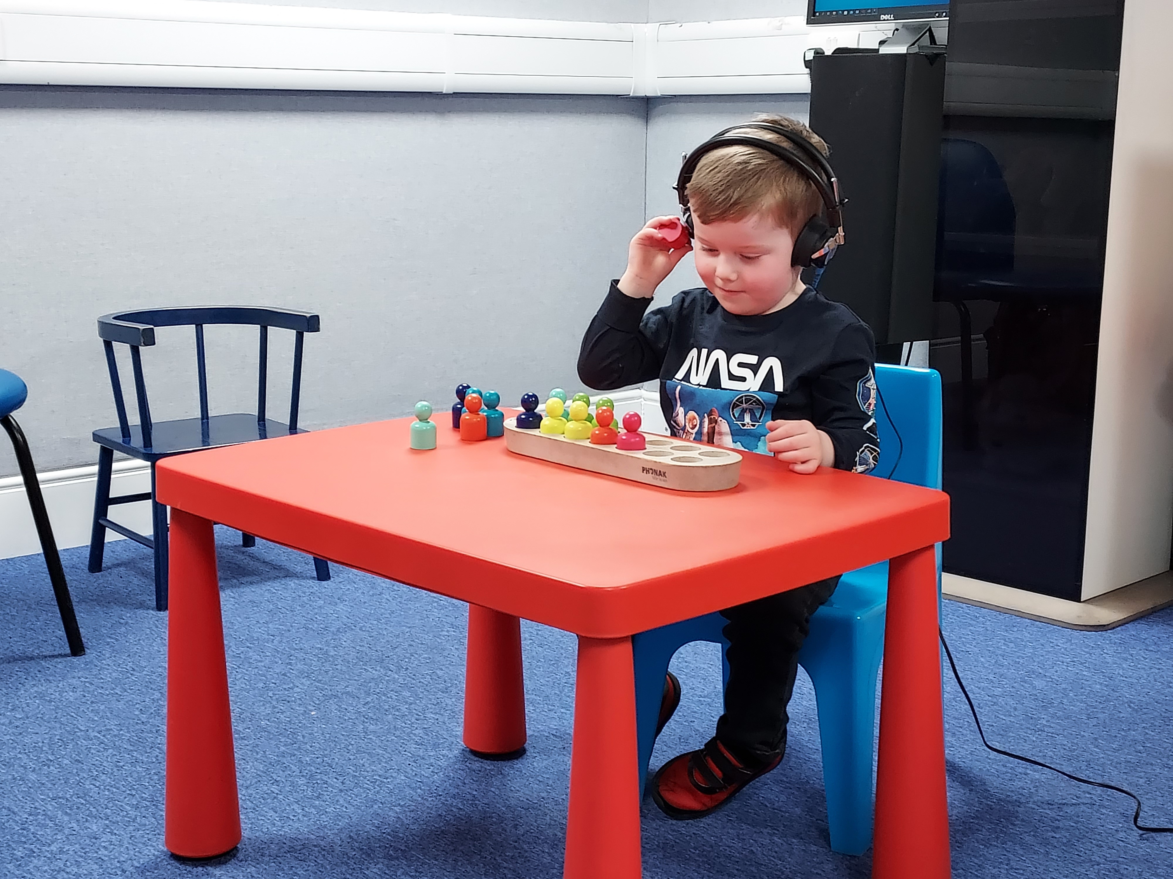 Young boy sat at a table wearing headphones during an audiology assessment, with coloured items in front of him.